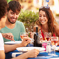 People dining outdoors, eating and laughing, seated at a table with wine glasses and colorful drinks.